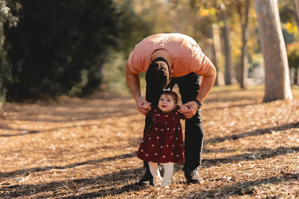 A parent helping a smiling toddler stand on a sunlit park path.