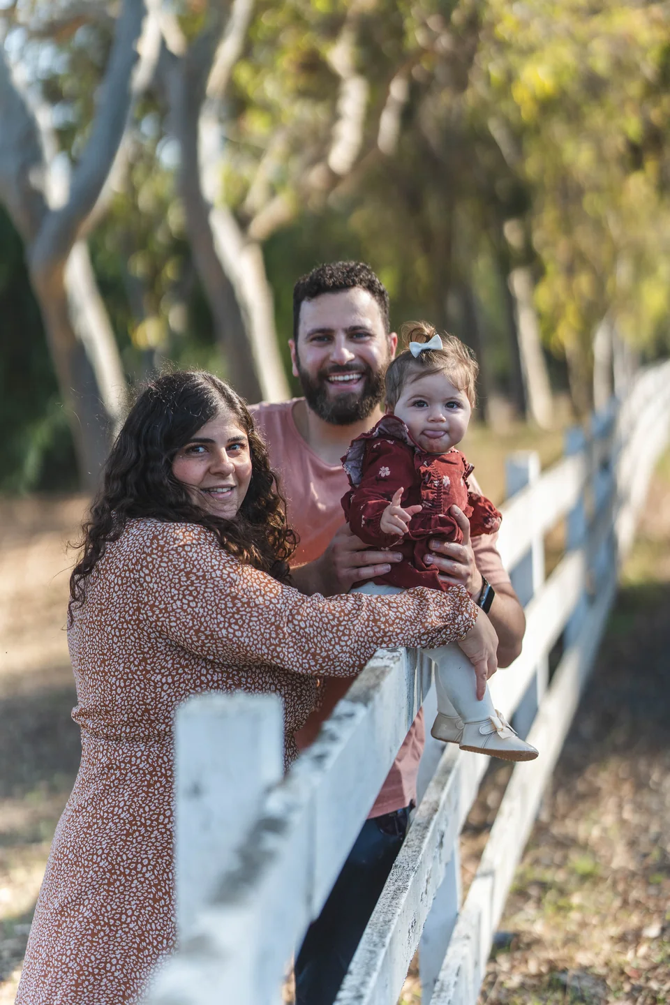 Two parents and their toddler leaning together beside a white fence in a park.