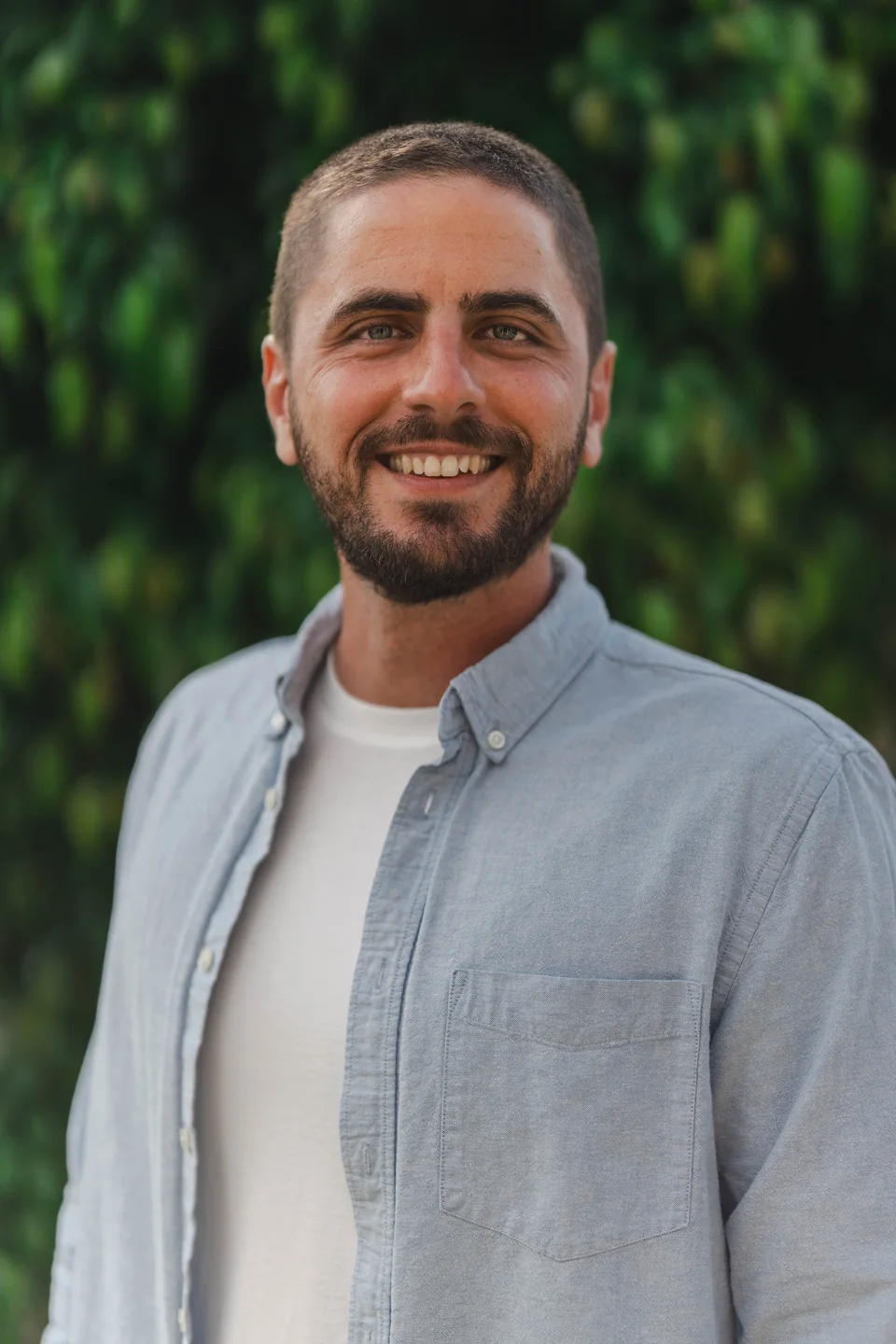 An outdoor headshot of a smiling man in a light blue shirt against soft green foliage.
