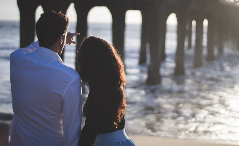 A couple seen from behind, looking toward the sunlit arches of a beach pier.
