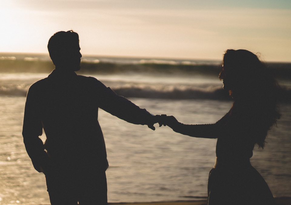 A couple holding hands in silhouette beside the waves.