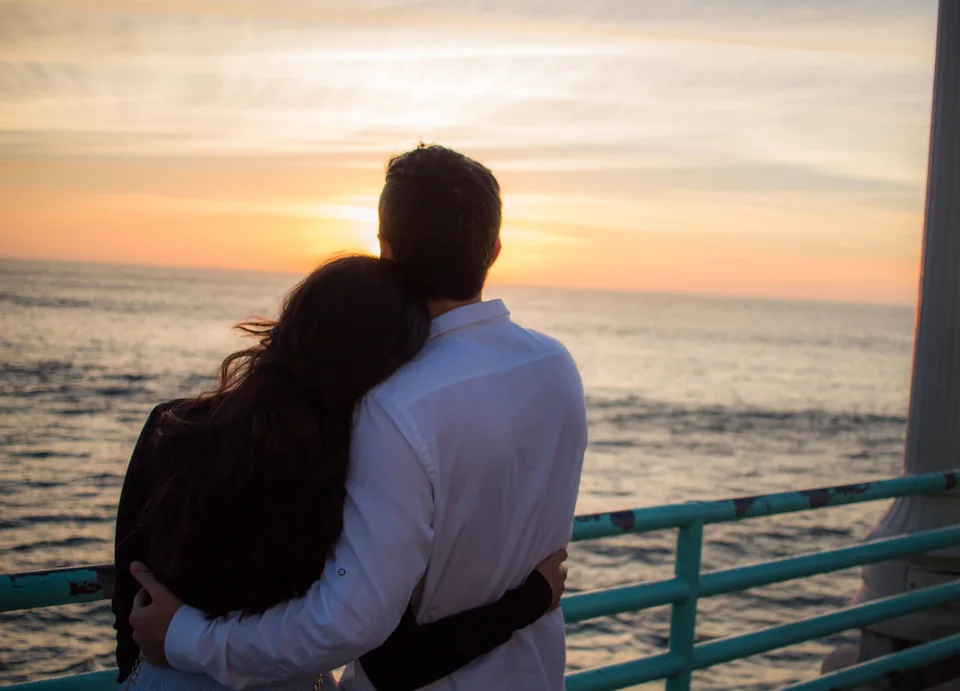 A couple embracing from behind as they watch the sunset over the ocean.