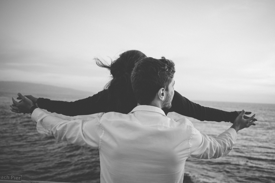 A black-and-white photograph of a couple opening their arms toward the ocean.