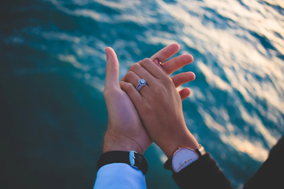 A couple holding hands beside the ocean, with an engagement ring visible.