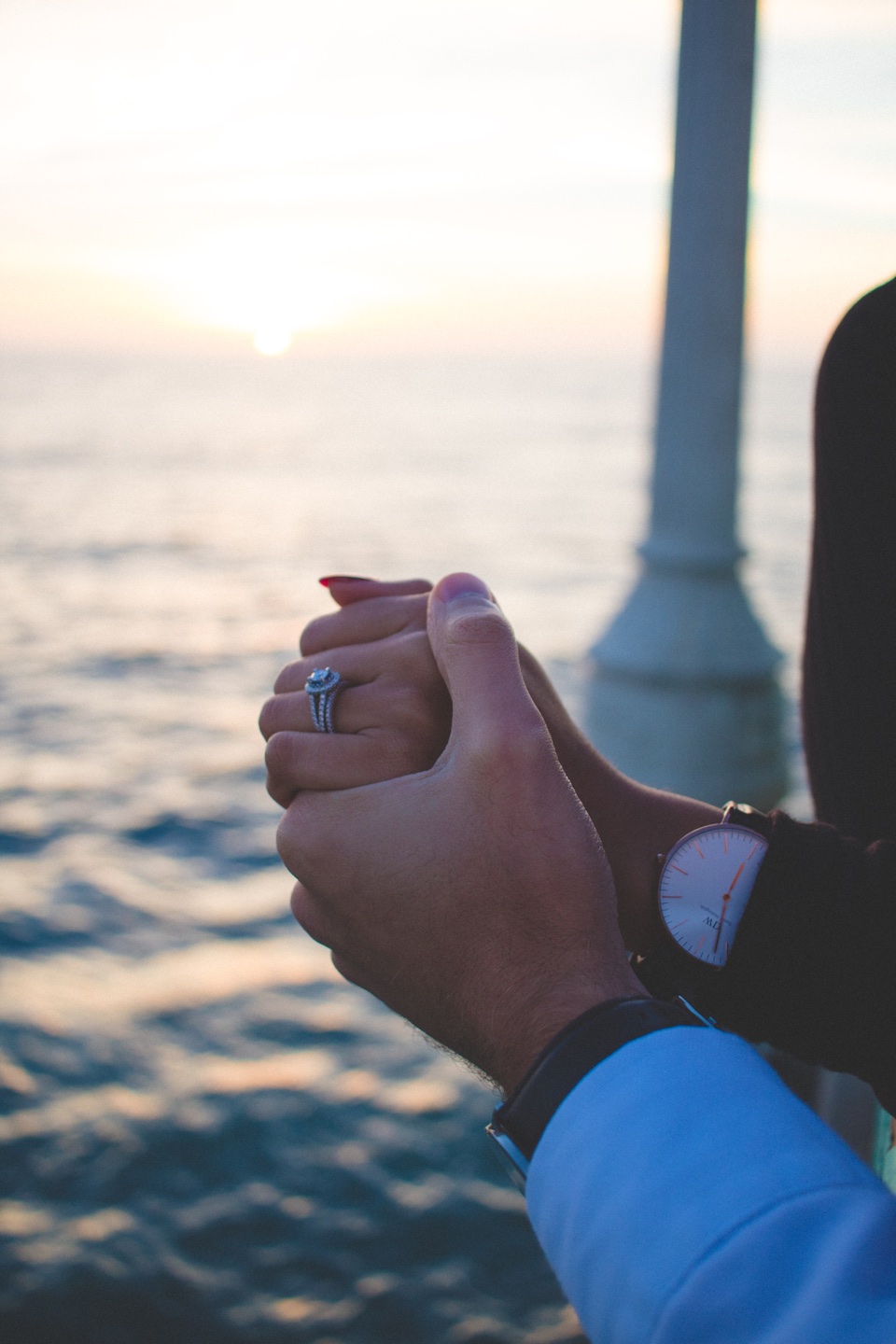 A close-up of clasped hands and an engagement ring at sunset.